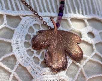 May include: A necklace featuring a copper-toned metal leaf pendant suspended from a delicate chain. The chain is accented with a strand of small, round, variegated purple and black beads that lead up to the metallic leaf-shaped centrepiece.
