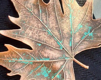 May include: A metallic maple leaf ornament featuring a copper-toned finish with visible teal-coloured oxidation accents. The leaf has a detailed, textured surface with prominent veins and jagged edges, showcasing a weathered, aged appearance.