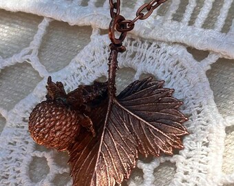 May include: A copper-toned pendant necklace featuring a detailed strawberry fruit and a single serrated leaf. The pendant is suspended from a matching copper-coloured metal chain link, showcasing a textured, metallic finish on both the fruit and leaf.