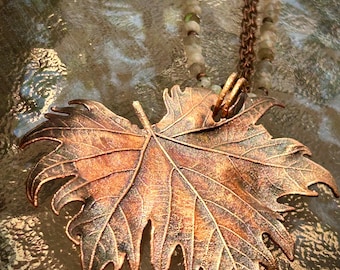 May include: A copper-toned metal pendant shaped like a maple leaf with visible veins and textured edges. The pendant hangs from a chain featuring small, multi-coloured, translucent beads and a dark metallic link chain, creating a layered necklace.