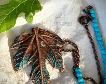 May include: A necklace featuring a large copper-toned leaf pendant with green patina accents and a single clear crystal centre. The chain is composed of a dark copper-coloured metal link section and a strand of round, bright turquoise blue beads.