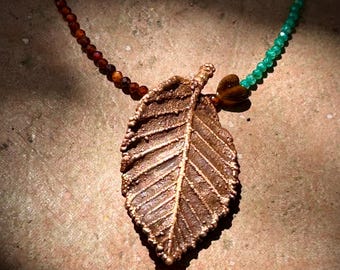 May include: A necklace featuring a central copper-coloured leaf pendant with a textured surface. The chain consists of small faceted beads, with dark reddish-brown beads on the left side and bright green beads on the right side, connected by a small heart-shaped bead.
