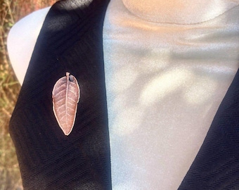May include: A copper-toned leaf-shaped brooch featuring a detailed vein pattern and a smooth, metallic finish. The brooch is pinned to a black sleeveless garment with a subtle textured chevron pattern, showcasing a clear stem at the top.