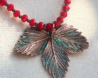 May include: A necklace featuring a large, copper-toned metal pendant shaped like a three-part leaf with oxidised teal green accents. The chain consists of faceted, bright red beads separated by small, dark metallic spacers and one turquoise bead.