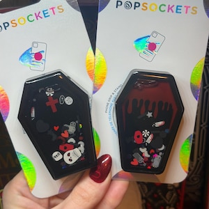 May include: Two coffin-shaped PopSockets phone grips and stands in retail packaging. The left grip features a black frame with a red cross, skull, and heart confetti. The right grip has a red interior with black drip accents and assorted confetti.