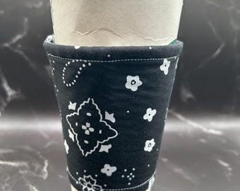 May include: A reusable coffee cup sleeve in black fabric with a white paisley and floral pattern. The sleeve is wrapped around a disposable paper coffee cup. The cup and sleeve are on a dark, marbled surface.