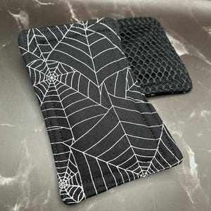 Op de afbeelding: Twee rechthoekige zwarte stoffen onderzetters. Eén heeft een wit spinnenwebontwerp, de andere een zwarte mesh textuur. Deze onderzetters zijn waarschijnlijk voor Halloween of griezelige decoraties.