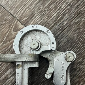 May include: A silver metal tube bender tool with degree markings from 45 to 180. The tool has a curved section and a handle with measurements. The text "1/2 O.D. 1 1/2 R." is visible.