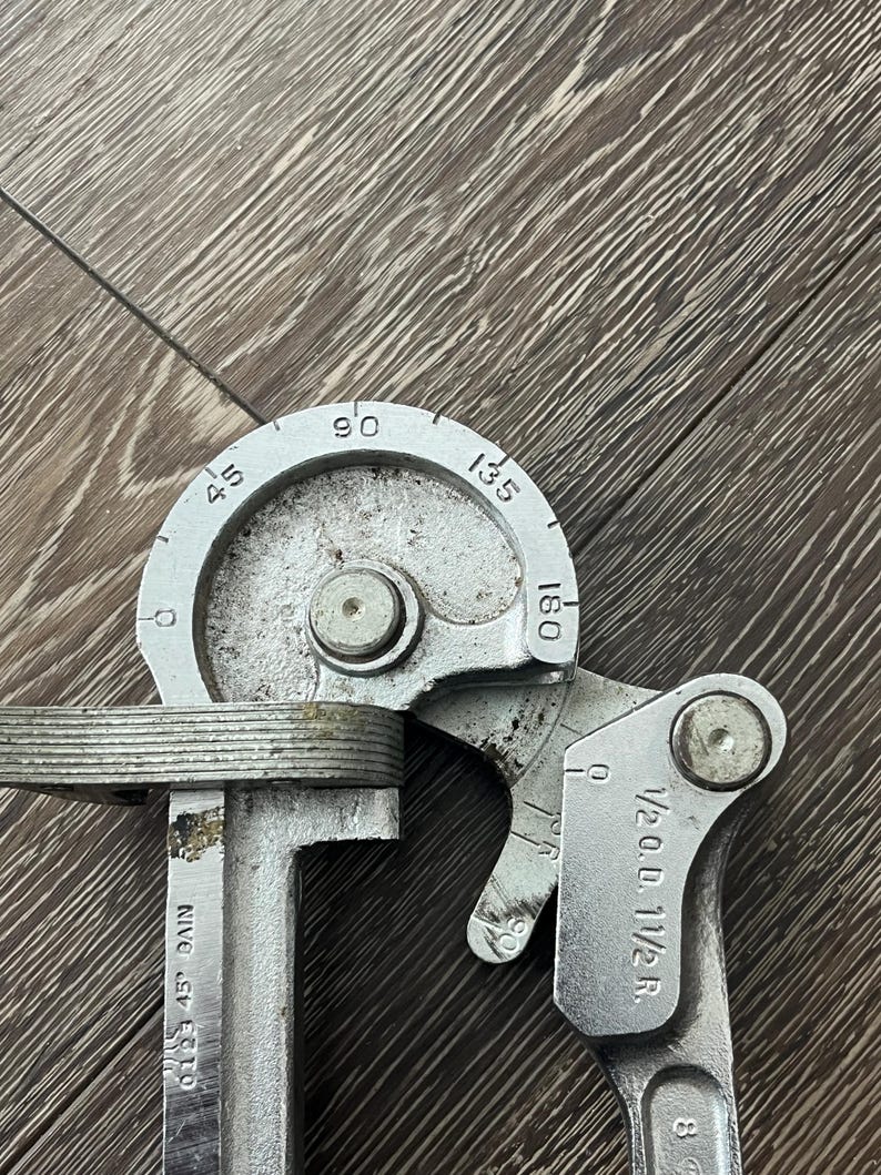 May include: A silver metal tube bender tool with degree markings from 45 to 180. The tool has a curved section and a handle with measurements. The text "1/2 O.D. 1 1/2 R." is visible.