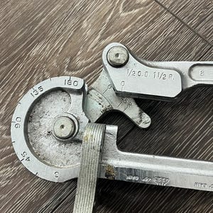 May include: A silver-colored pipe bender tool with measurements for angles from 45 to 180 degrees. The tool has markings including "1/2 O.D. 1 1/2 R." and "0123 45 CAM". The tool is designed for bending pipes.