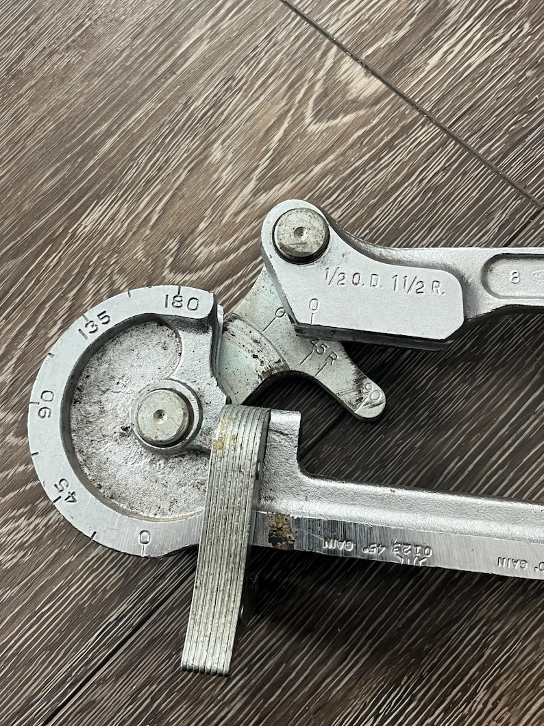 May include: A silver-colored pipe bender tool with measurements for angles from 45 to 180 degrees. The tool has markings including "1/2 O.D. 1 1/2 R." and "0123 45 CAM". The tool is designed for bending pipes.