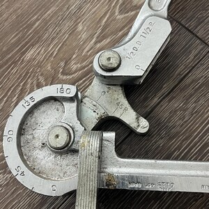 May include: A silver-colored pipe bender tool with degree markings from 0 to 180. The tool has a handle and a curved section for bending pipes. Markings include "1/2 O.D. 11/2 R." and "0123 45 CAIN".