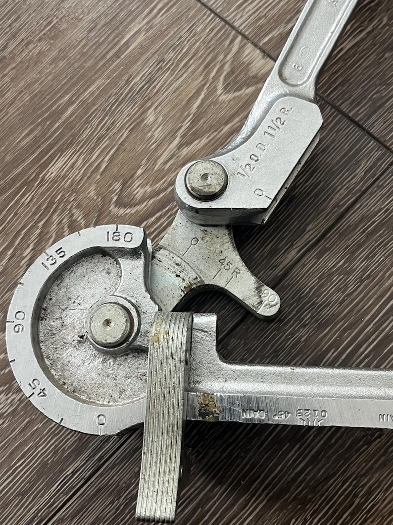 May include: A silver-colored pipe bender tool with degree markings from 0 to 180. The tool has a handle and a curved section for bending pipes. Markings include "1/2 O.D. 11/2 R." and "0123 45 CAIN".