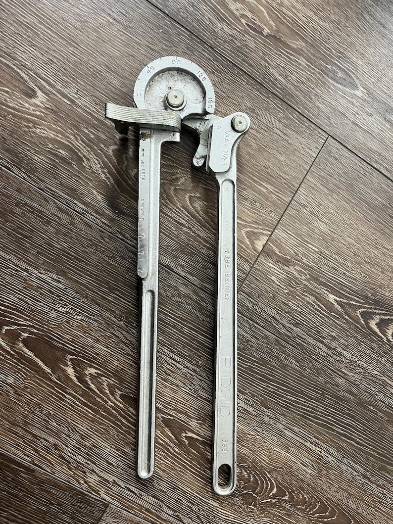 May include: A silver-colored tube bender tool with a curved head and a long handle. The tool has markings for various bending angles, including 45, 90, 135, and 180 degrees. The handle is marked "RIDGID" and "398".