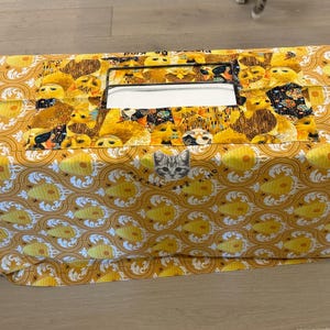 May include: A rectangular tissue box cover with a yellow and white patterned fabric. The top features a rectangular opening for tissue access, and a section with cat-themed fabric. A small cat face is visible.