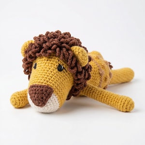 Puede incluir: Un juguete de león de ganchillo hecho a mano en tonos de amarillo, marrón y crema. El león presenta una melena marrón detallada, ojos negros pequeños y un hocico color crema. El juguete está tumbado, con las patas extendidas.