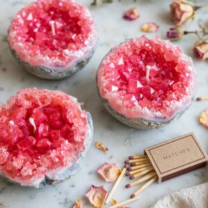 May include: Three round candles with a textured grey exterior and a vibrant pink crystalline interior featuring a central wick. A small rectangular cardboard box labelled MATCHES with several wooden matches scattered nearby is also present.