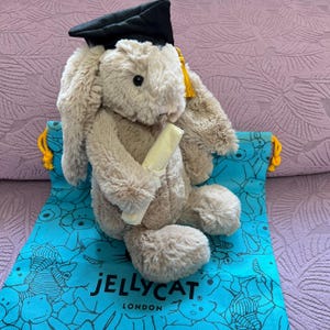 Puede incluir: Un conejo de peluche marrón claro con un gorro de graduación negro con borla dorada, sosteniendo un diploma enrollado. El conejo está sobre una bolsa azul con un patrón de osos y el texto "JELLYCAT LONDON".