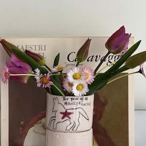 Peut inclure: Un vase cylindrique en céramique blanche orné d'un dessin au trait noir représentant un cheval avec une étoile rouge sur le corps et l'inscription "The year of a lucky horse!", rempli de tulipes violettes et de marguerites blanches.