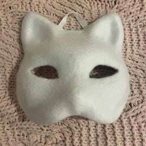 May include: A white cat mask with eye cutouts and a white ribbon for securing. The mask is made of a soft, felt-like material and is designed to cover the upper face. The mask is set against a textured, pink background.