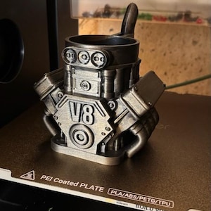 May include: A metallic silver-colored 3D printed mug shaped like a V8 engine block. The front of the mug features the embossed text V8, along with circular details resembling engine components and a handle extending from the back side.