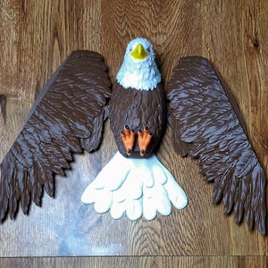 May include: A bald eagle figurine featuring a white head and tail, brown wings and body, a yellow beak, and orange talons. The eagle is depicted with its wings spread wide in a flat, horizontal position, showcasing detailed molded feather textures.
