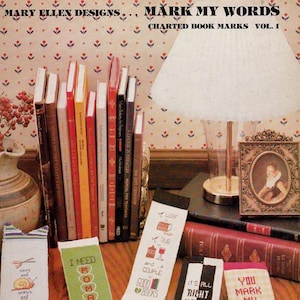 May include: A collection of five rectangular cross-stitch bookmarks featuring various embroidered designs and text including I NEED ROMANCE, IT'S ALL RIGHT HERE IN BLACK AND WHITE, and YOU MARK MY WORDS I SHALL RETURN, displayed on a wooden surface.