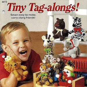 May include: A craft pattern booklet titled Tiny Tag-alongs! featuring seven crocheted animal figures including a lion, bear, zebra, giraffe, elephant, monkey, tiger, and a clown. The cover displays the finished yarn toys arranged on a wooden bench.