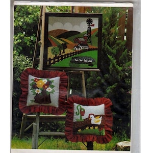 May include: A package for a Fantasy Farm applique pattern set. The set includes patterns and instructions for a 20" x 20" wall hanging and two 12" x 12" ruffled pillows. The image shows the finished products: a framed farm scene and two pillows with floral and farm animal designs.