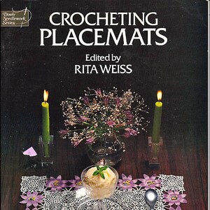 May include: Crocheting Placemats, a book with a crocheted placemat on the cover, featuring a white crocheted placemat with purple flowers, a glass dish with a scoop of ice cream, and two lit candles.
