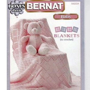May include: A pink crocheted baby blanket with a teddy bear sitting on top of it. The blanket is on a white chair. The image is from a Bernat crochet pattern book. The text on the cover reads "Baby Blankets (to crochet)"