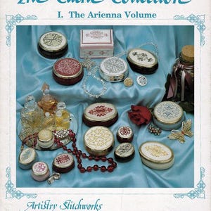 May include: A vintage book cover titled "The Cache Collection I. The Arienna Volume" featuring various decorative boxes with embroidered designs in red, white, and gold. The cover also includes the text "Artistry Stitchworks".