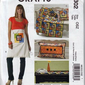 May include: A McCall's Crafts sewing pattern, M6302, featuring a woman wearing an apron. The pattern includes instructions for a quilt, pillows, and a table runner with colorful square designs. The apron has a colorful square design.