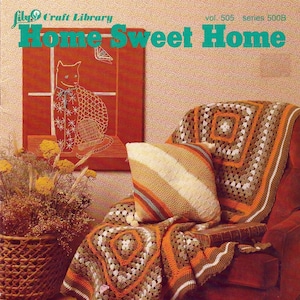 May include: A crocheted throw blanket in shades of brown, orange, and white is draped over a red armchair. The blanket has a square pattern with a smaller square in the center. A pillow with brown and orange stripes is on the chair. The image is from a craft book titled "Home Sweet Home" with the volume and series numbers.