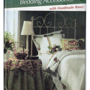 May include: A Simplicity Home Decorating pattern for bedding accessories with handmade roses. The pattern number is 4569. The image shows a white bed with a white bedspread and a white pillow with a teddy bear on it. There is a green table with a floral tablecloth and a lamp on the table.