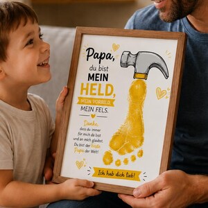 K&ouml;nnte beinhalten: Ein gerahmter Druck mit dem Text "Papa, du bist MEIN HELD" und einem gelben Fu&szlig;abdruck-Design. Der Druck enth&auml;lt auch den Text "Ich hab dich lieb!" und wird von einem Mann und einem Kind gehalten.