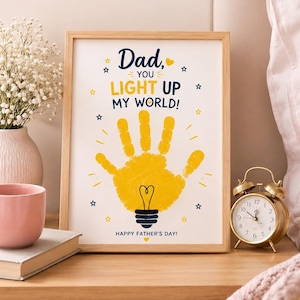 May include: A framed print with the text "Dad, you light up my world!" and a yellow handprint shaped like a lightbulb. The print is on a wooden nightstand with a pink mug, a book, and a vase of white flowers.