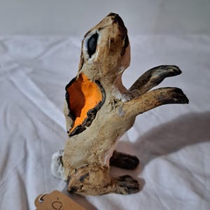 May include: A tan and brown ceramic rabbit figurine in a standing pose with its front paws raised. The rabbit features a bright orange interior on one ear, dark brown accents on the paws and face, and a small white tail. A brown tag reads Clover.