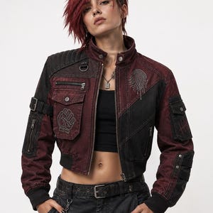 Puede incluir: Una chaqueta bomber corta en negro y rojo oscuro con cuello alto, bolsillos con cremallera y correas con hebilla en las mangas. El pecho muestra un gráfico de una huella dactilar y un puño, con un tejido de acabado desgastado.