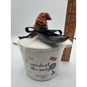 May include: A white ceramic canister with a brown Sorting Hat lid topper, featuring the text wands at the ready and various small icons. A black ribbon and a Harry Potter Rae Dunn tag are attached. A wooden ruler shows a height of 7 inches.