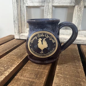 May include: A dark blue ceramic mug with a glossy finish and a large handle. The front features a circular beige emblem with a rooster illustration and the text Sunny Side Up Grill, Where strangers Become friends, LITTLE RIVER, SC.