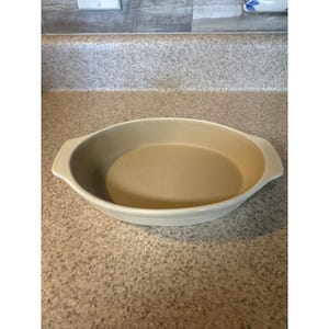 May include: An oval-shaped ceramic baking dish with a smooth, light beige interior and a slightly darker beige exterior. The dish features two integrated handles on opposite sides and a shallow, wide basin design for cooking or serving food.