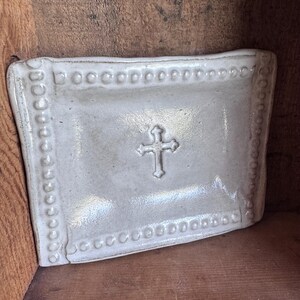 May include: A rectangular, off-white ceramic dish with a raised cross design in the center. The edges are adorned with a series of small, raised dots. The dish has a slightly curved shape.