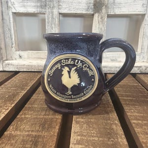 May include: A dark blue ceramic mug with a glossy finish and a rounded handle. The front features a circular cream-colored emblem with a rooster illustration and the text Sunny Side Up Grill, where strangers become friends, LITTLE RIVER, SC.