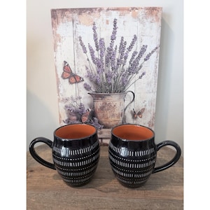 May include: Two black ceramic mugs with a rounded shape and a burnt orange interior. The exterior of each mug features a repeating pattern of vertical white dashes arranged in horizontal bands around the body and a curved handle on the side.