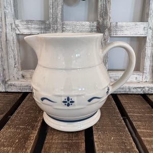 Op de afbeelding: Een crèmekleurige keramische kan met een ronde vorm, een gebogen handvat en een schenktuit. De buitenkant is versierd met een herhalend patroon van kleine blauwe bloemmotieven en horizontale blauwe lijnen rond de basis en het midden.