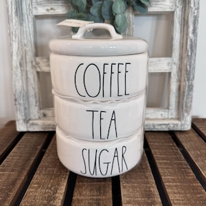 May include: A white ceramic canister set with three stacked containers. Each container is labeled in black script: "COFFEE", "TEA", and "SUGAR". The set has a matching lid with a handle. The background includes a wooden surface and a decorative window.