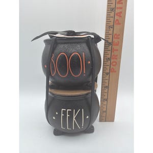 May include: A stacked set of two black ceramic cauldrons held by a black metal frame. The top cauldron features the orange text BOO! with small stars, and the bottom cauldron features the white text EEK! with stars. A wooden ruler shows a height of 8 inches.