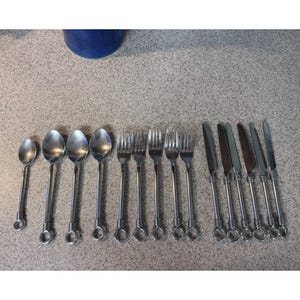 May include: A collection of stainless steel silverware, including spoons, forks, and knives. The silverware has a decorative handle design with a ring detail. The arrangement is on a neutral-colored surface.