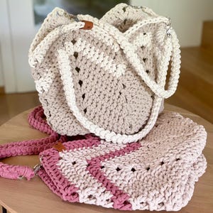 Könnte beinhalten: Zwei handgefertigte Häkel-Tragetaschen mit dickem geflochtenem Riemen und strukturiertem Zickzackmuster. Eine Tasche ist aus cremefarbenem und beigem Garn gefertigt, die andere in hellen und dunklen Rosatönen, beide mit silbernen Metallbeschlägen.